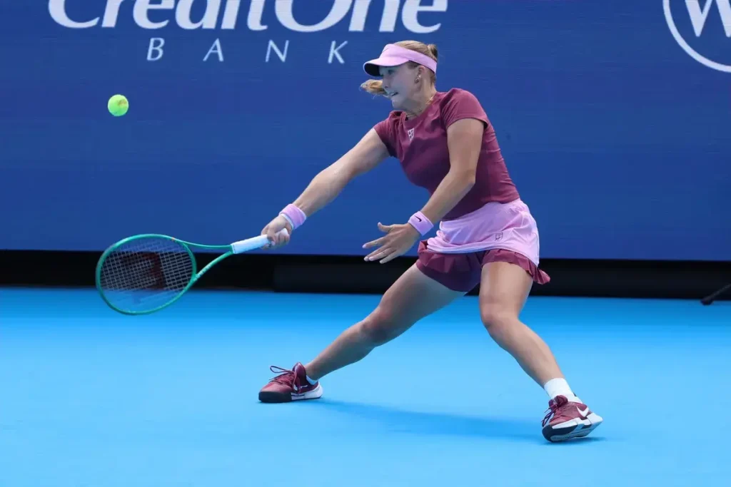Tennis player in maroon top and pink visor hits a backhand on a blue court, Credit One Bank sponsor board behind her