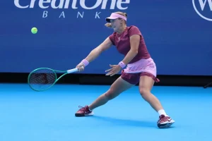 Tennis player in maroon top and pink visor hits a backhand on a blue court, Credit One Bank sponsor board behind her