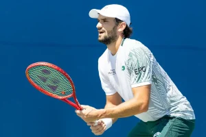 Tennis player in white cap and green-and-white kit holding a red Yonex racket on a blue court, preparing to strike a return.
