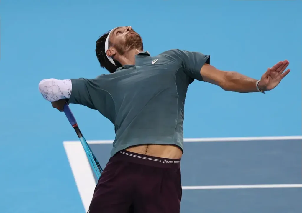 Tennis player in grey Asics shirt and maroon shorts, arms outstretched and head tilted back, holding a racket with a taped wrist on a blue court