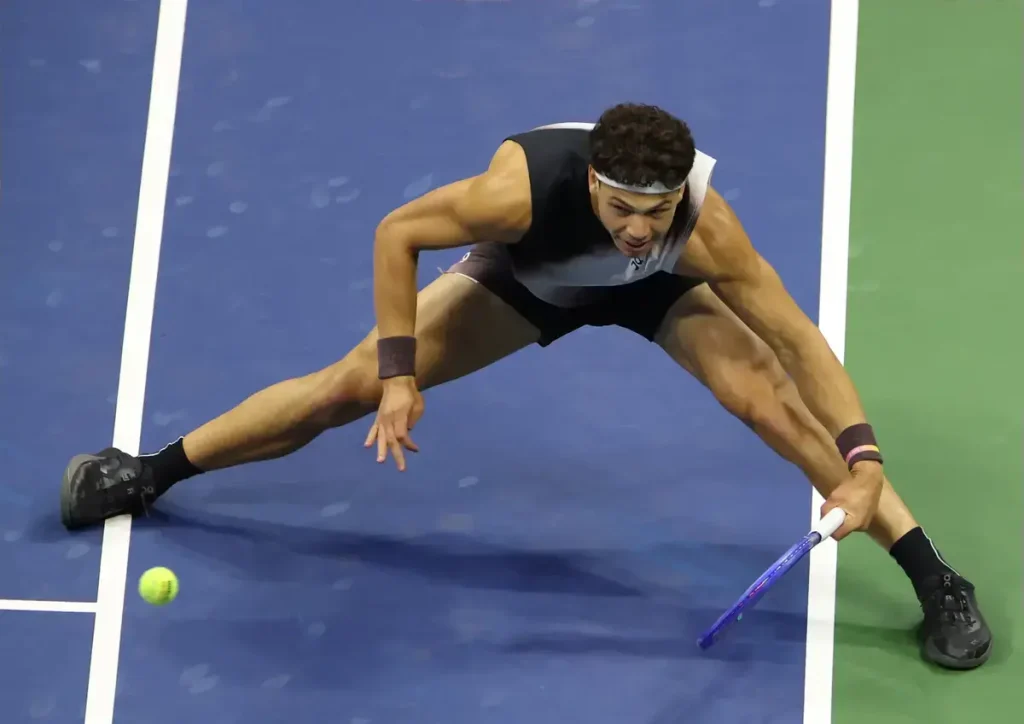 Tennis player in black sleeveless top and headband stretching low to reach a yellow ball on a blue court near the sideline.