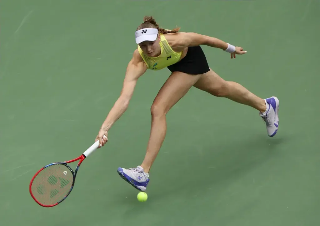 Elena Rybakina in yellow top and white visor reaches low for a backhand on a green hard court at a tennis match.