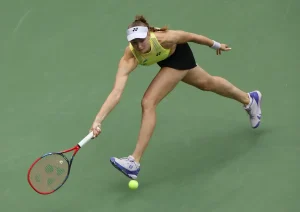 Elena Rybakina in yellow top and white visor reaches low for a backhand on a green hard court at a tennis match.