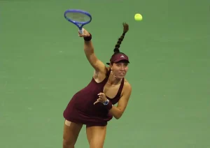 Tennis player in a maroon visor and dress, braided ponytail, serving with a blue racket on a green court at night.