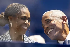 Two smiling people, one with braided hair in a bun and striped jacket, the other with grey hair and a blue tie, share a close moment at an outdoor event.