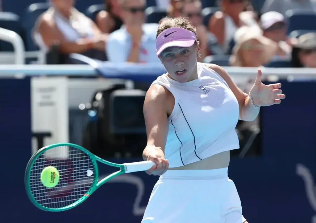Tennis player in a lilac visor and white outfit hitting a forehand with a green Wilson racquet at a court. Name not visible.
