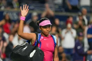 Tennis player in a pink visor, pink top and wristband waves to fans while carrying a black Nike bag at a night match.