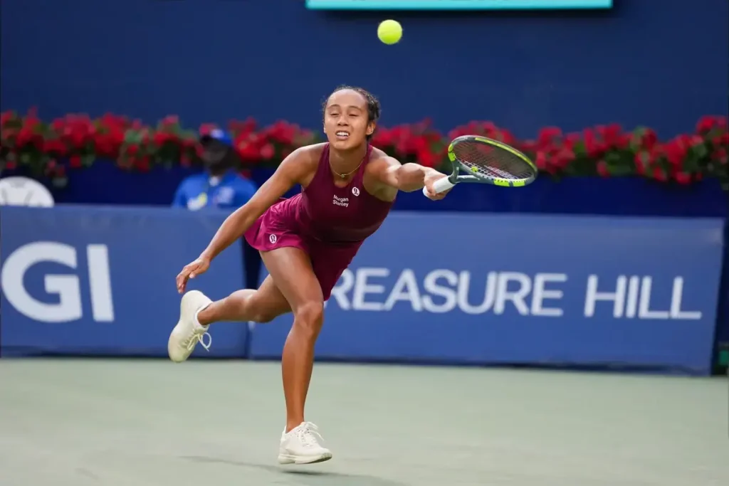 Leylah Fernandez in a maroon outfit lunges for a low forehand return on a hard court at the Singapore Open, with flowers and sponsor signage behind her.