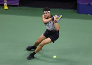 Ben Shelton hitting a backhand on a green hard court, wearing a white headband, dark outfit and a purple-accented racket, mid-rally.