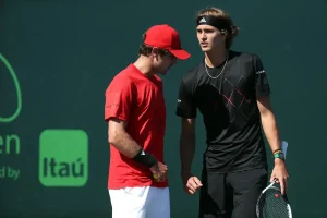 Two tennis players in conversation on a court; one in a red cap and red shirt, one in black with a headband and racket. Green Itaú sponsor board behind them.
