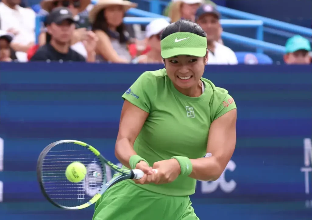 A tennis player in a lime green visor, top and wristbands hits a forehand on court, with spectators behind her at the tournament.