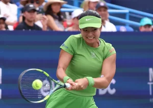 A tennis player in a lime green visor, top and wristbands hits a forehand on court, with spectators behind her at the tournament.