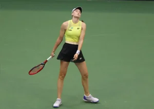 Tennis player in yellow Yonex top and black skirt holding a red racquet, head tilted back in frustration on a green indoor court