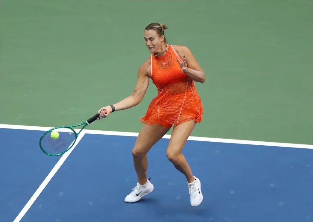 Tennis player in an orange mesh dress swinging a green racquet to return a ball on a blue court