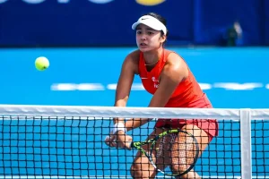 Alex Eala in a red Indonesia-branded top and white visor returning a shot at the net during a match on a blue hard court.
