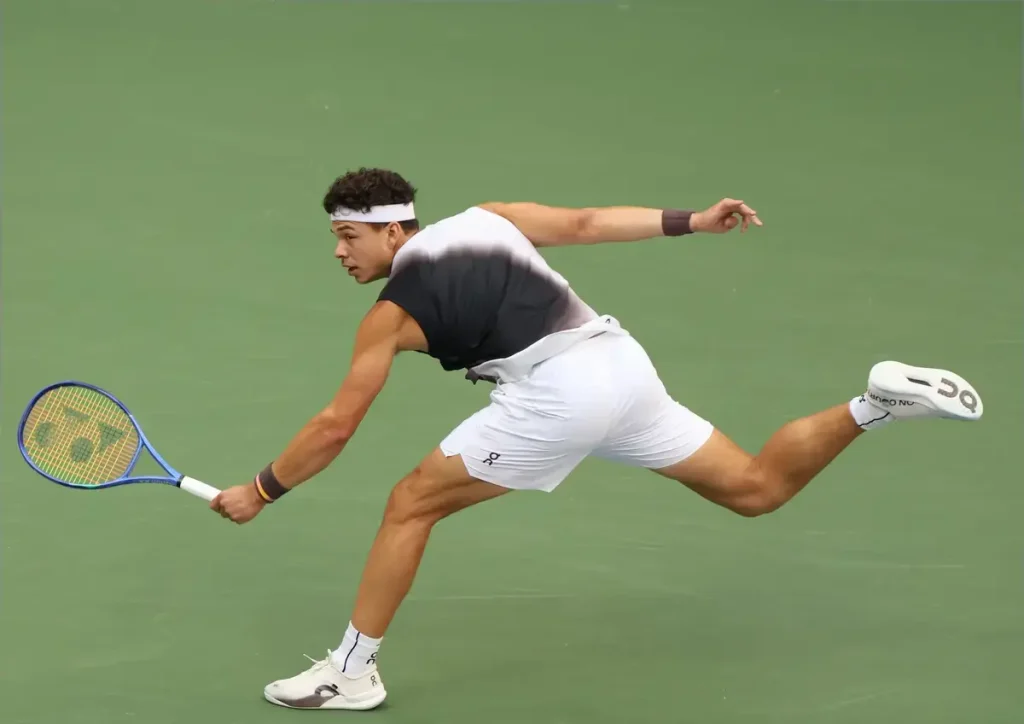 Tennis player in white shorts and black shirt, reaching low for a forehand with a Yonex racquet on a green court at the US Open