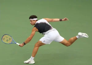 Tennis player in white shorts and black shirt, reaching low for a forehand with a Yonex racquet on a green court at the US Open