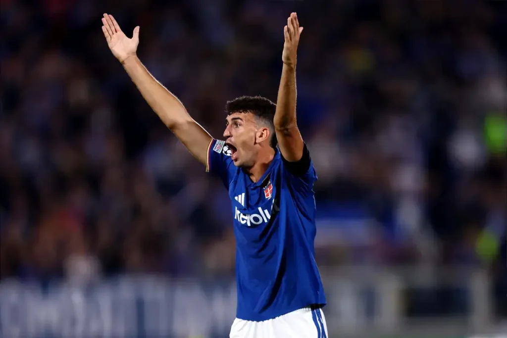 Jacobo Ramon in blue Real Oviedo kit, arms raised and shouting at a match, with a blurred stadium crowd behind him.