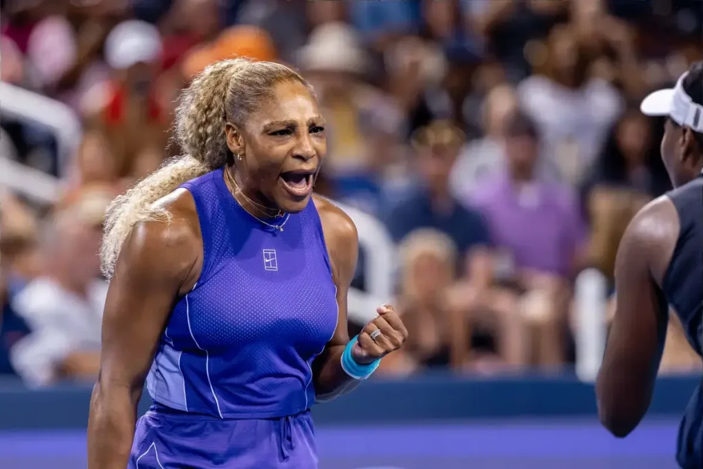 A tennis player in a purple sleeveless top and visor shouts and clenches her fist during a match, with a blurred crowd behind her and an opponent in the fore…