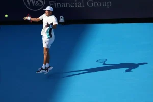 A tennis player in white cap and kit mid-forehand on a blue court beside a "W&S Financial Group" banner, his long shadow stretched across the court.
