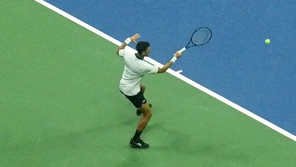 Tennis player in white shirt and black shorts hitting a forehand on a green-and-blue hard court, ball in the air beside him.