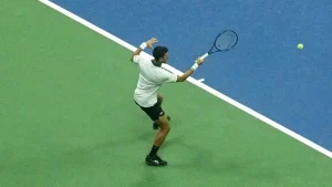 Tennis player in white shirt and black shorts hitting a forehand on a green-and-blue hard court, ball in the air beside him.