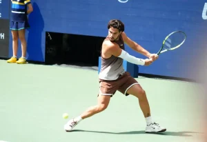 Tennis player in brown outfit and white sleeve hits a forehand on a hard court, with a ball and courtside ball kid visible.