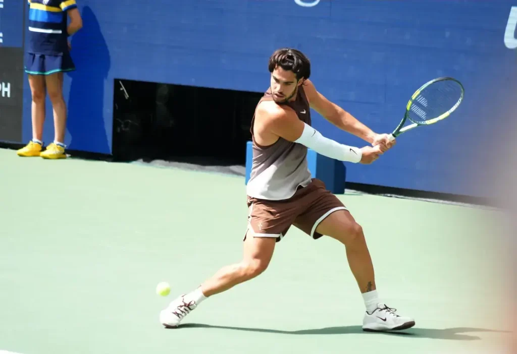 Tennis player in brown outfit and white sleeve hits a forehand on a hard court, with a ball and courtside ball kid visible.