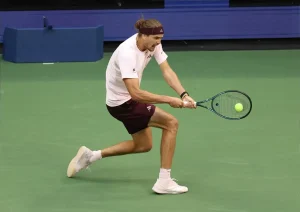 Alexander Zverev in maroon shorts and white shirt lunging to hit a forehand on a green hard court at a tennis tournament.