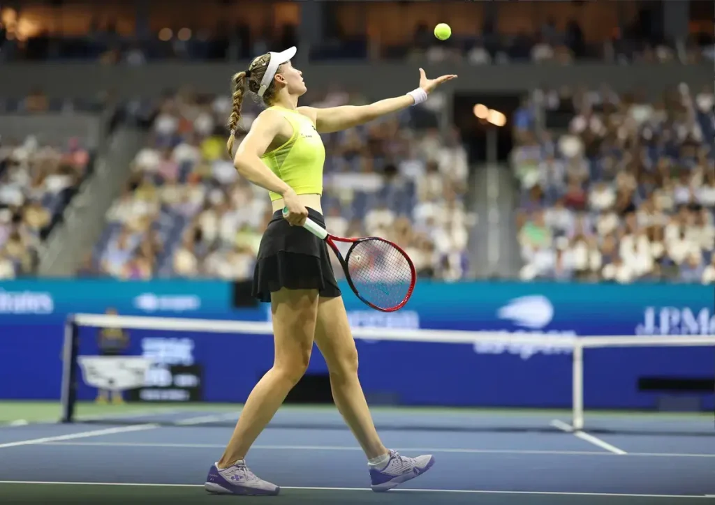 Tennis player in yellow top and black skirt tossing a ball before serving at a packed stadium, with blue court and sponsor banners behind her.