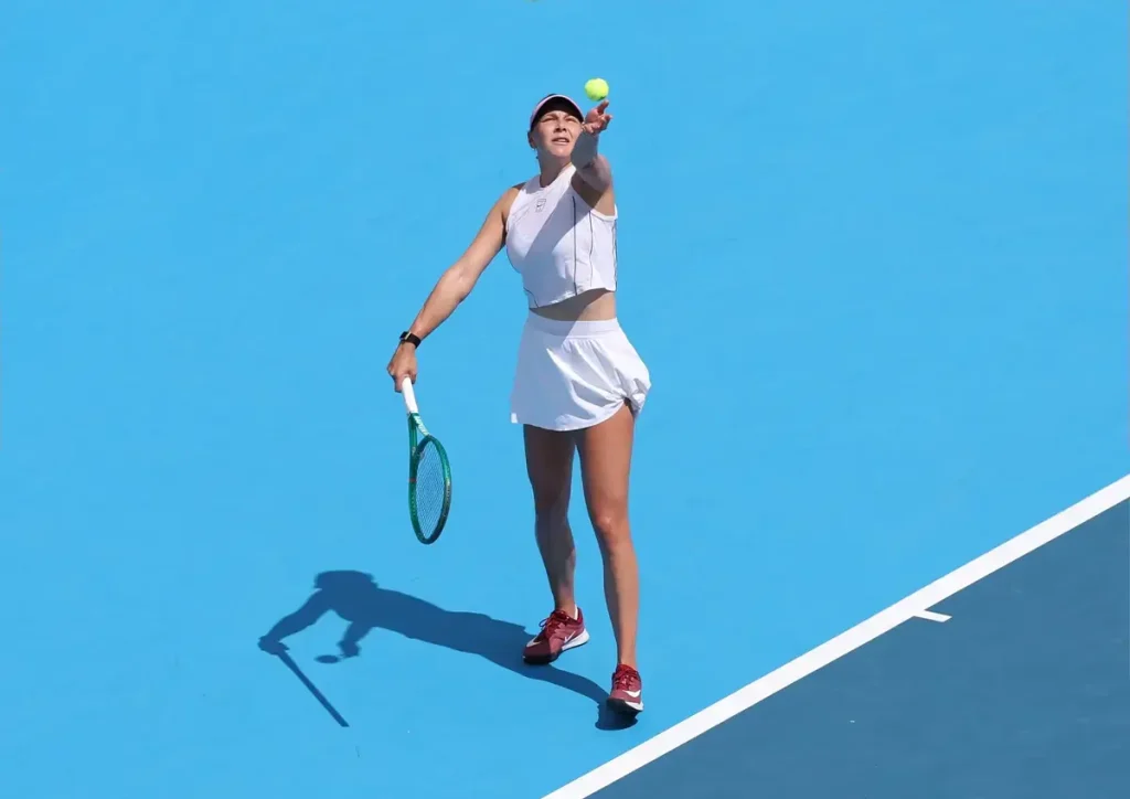 A tennis player in a white outfit and visor serves on a bright blue court, tossing the ball overhead with a green racket in hand