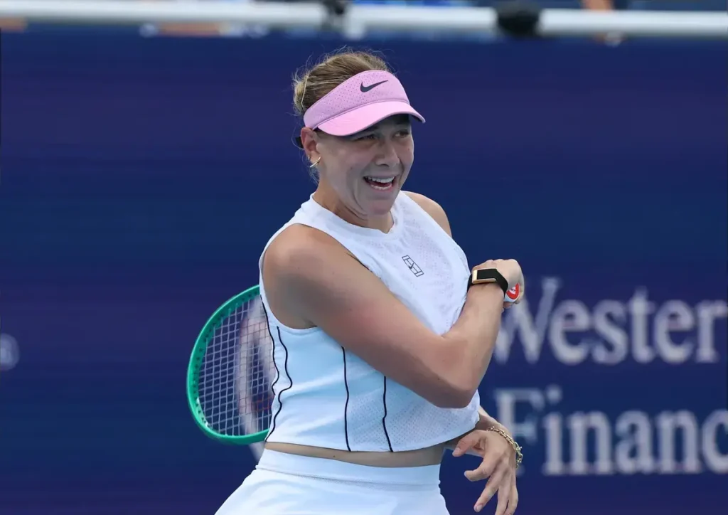 Amanda Anisimova smiling on court in a white outfit and pink visor, holding her racket and checking her wrist watch at a tournament