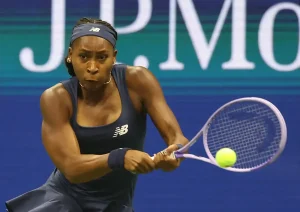 Tennis player in navy New Balance kit and headband hitting a forehand with a purple racket, blurred "J.P. Morgan" signage behind her at a tournament.