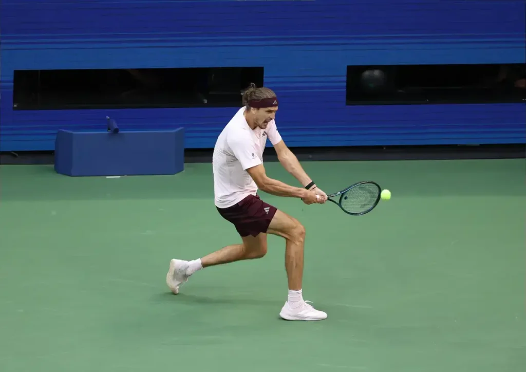 A tennis player in a white shirt and maroon shorts hits a backhand on a green court, with a yellow ball near his racket.