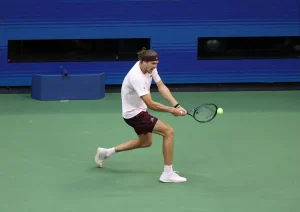 A tennis player in a white shirt and maroon shorts hits a backhand on a green court, with a yellow ball near his racket.