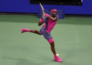Frances Tiafoe in pink and grey hitting a forehand on a green hard court during a match.