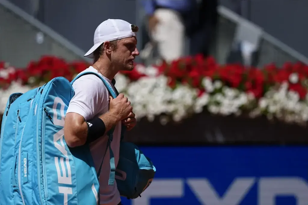 Tennis player in white cap and white shirt carrying a blue Head racket bag, walking past red and white flowers at a clay court tournament