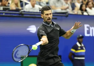 Tennis player in black shirt hits a forehand at a blue-court Grand Slam match, with spectators and a ball kid in the background