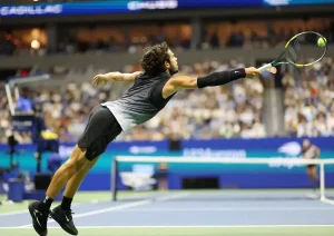 Tennis player in black sleeveless top and shorts stretching low to reach a ball with a racket at a night match, crowd behind