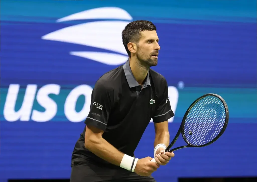 Tennis player in black Lacoste polo with Qatar Airways sponsor, holding a Head racquet on court in front of a US Open backdrop, looking focused during a match.