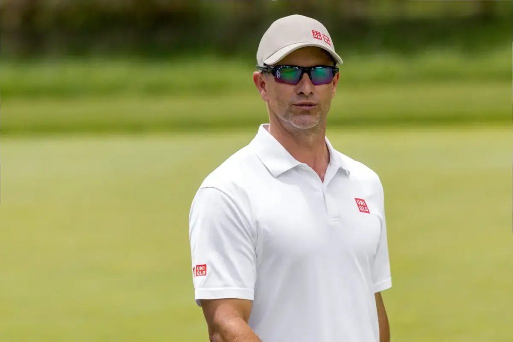 Man in white Uniqlo polo, beige cap and mirrored sunglasses, standing on a golf course green; the article concerns Adam Scott.