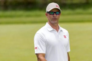 Man in white Uniqlo polo, beige cap and mirrored sunglasses, standing on a golf course green; the article concerns Adam Scott.