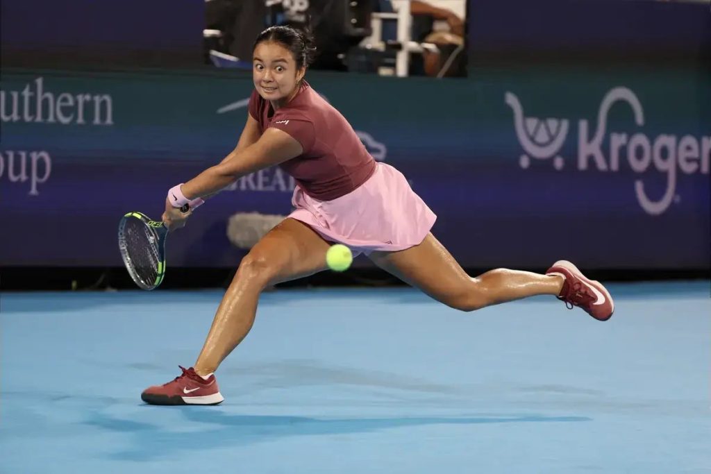 Tennis player in maroon top and pink skirt lunging to hit a low forehand on a blue hard court, with a Kroger sign behind her.