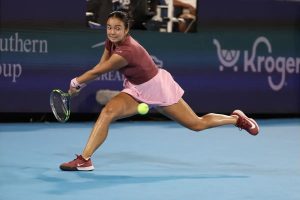 Tennis player in maroon top and pink skirt lunging to hit a low forehand on a blue hard court, with a Kroger sign behind her.