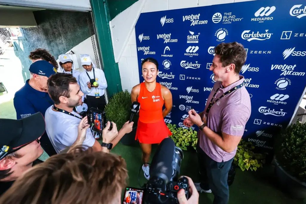 Woman in orange tennis dress speaks to reporters in front of a blue sponsor backdrop at a tournament, with cameras and microphones pointed at her