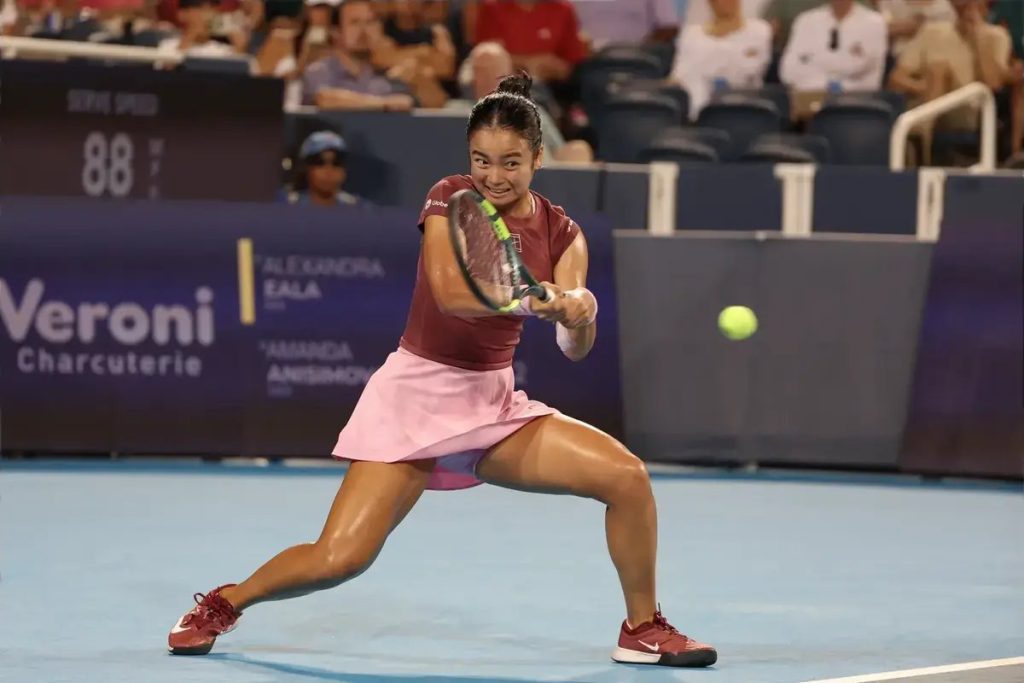 Alex Eala in a maroon top and pink skirt hits a forehand on a blue court during a night match, with a Veroni Charcuterie sponsor board behind her.