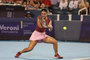 Alex Eala in a maroon top and pink skirt hits a forehand on a blue court during a night match, with a Veroni Charcuterie sponsor board behind her.