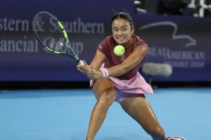 Tennis player in maroon top and pink skirt lunges to hit a backhand, ball in view, at a Western & Southern Open court with blue surface.