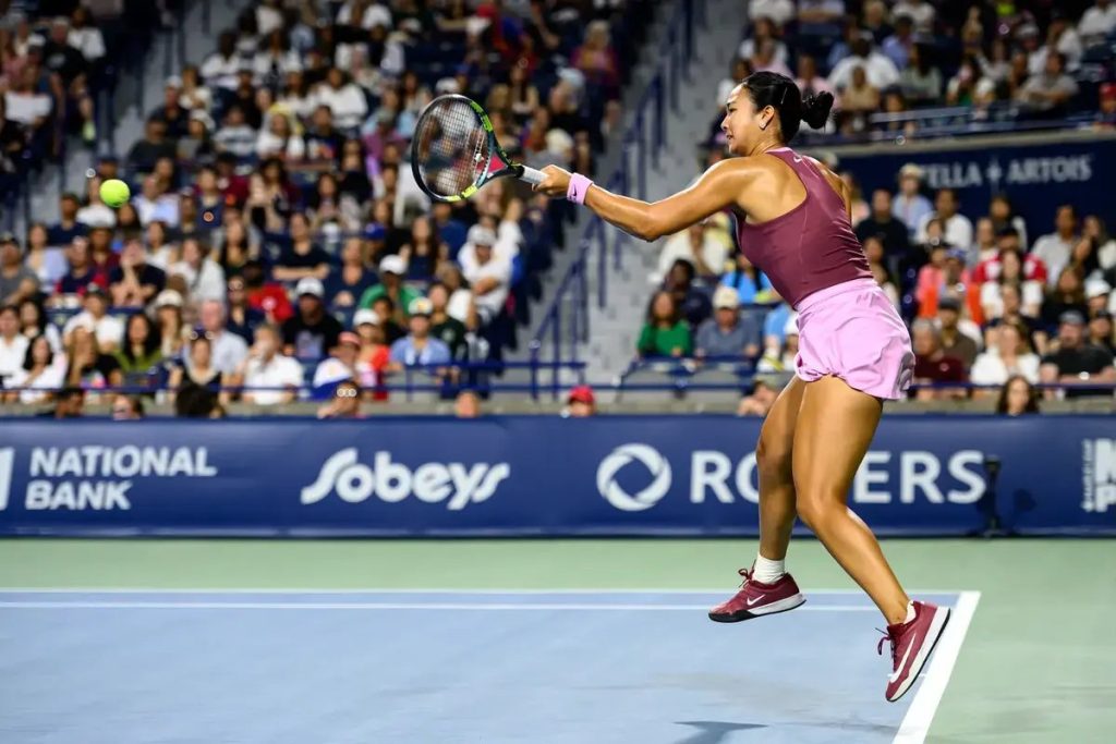 Alex Eala hits a forehand return in a purple outfit at a night tennis match, with a packed crowd behind her.