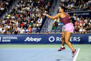Alex Eala hits a forehand return in a purple outfit at a night tennis match, with a packed crowd behind her.
